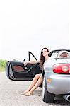 Young woman sitting in convertible on country road against clear sky