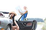 Woman using cell phone in convertible while friend reading map on road