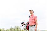 Thoughtful middle-aged golfer looking away while carrying bag against clear sky