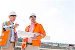 Supervisors with blueprints discussing at construction site against clear sky