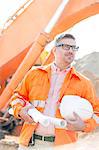 Thoughtful architect holding hardhat and blueprints at construction site