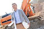 Smiling architect looking away while holding blueprints and hardhat at construction site