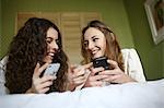 2 teens playing with their mobile phone on a bed