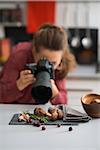 A woman photographer is leaning down, taking a close-up of autumn fruits and vegetables - mushrooms, garlic, rosemary, and cranberries. A wooden bowl sits on the modern kitchen counter.