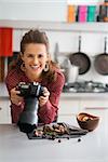 A woman food photographer looks up from her camera, smiling and happy. She is in a modern kitchen, photographing autumn fruits and vegetables - mushrooms, garlic, rosemary, and cranberries.