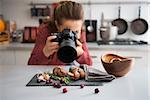 A woman food photographer in the background leans down to take a close-up, in a modern kitchen, of autumn fruits and vegetables - mushrooms, garlic, rosemary, and cranberries.