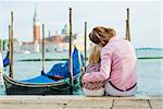 An elegant mother is sitting next to her daughter along the water's edge near St. Mark's Square in Venice. She is hugging her daughter close, resting her head against her daughter's head.