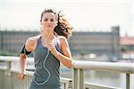 An athletic woman is jogging on a bridge, listening to music. Her long hair is up in a ponytail and blowing in the wind.