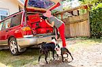 Woman packs luggage into the trunk of a car while her dogs watch