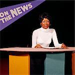 1970s AFRICAN AMERICAN TV ANCHORWOMAN AT BROADCAST DESK WITH STATION PROGRAM BANNER BEHIND