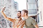 Male couple standing on balcony, taking self portrait, using smartphone