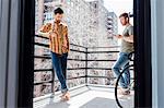 Male couple standing on balcony, standing away from each  other, drinking wine and looking at smartphone