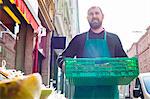 Grocer carrying food in crate by greengrocer's shop