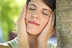 Woman leaning against tree trunk with hands on cheeks, eyes closed