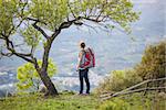Woman hiker with backpack standing under tree and enjoying valley view