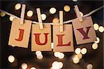The word JULY printed on clothespin clipped cards in front of defocused glowing lights.