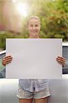 Pretty Young Woman Holding a Clean White Board with Copy Space and Smiling at the Camera in front of her Car