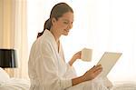 Woman in bathrobe drinking coffee and using digital tablet in bedroom