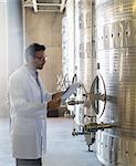 Vintner in lab coat with clipboard checking stainless steel vat in winery cellar
