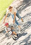 Mother pushing son with helmet on bicycle in sunny park