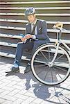 Businessman in suit and helmet texting with cell phone next to bicycle on sunny urban stairs