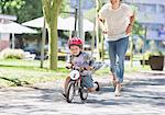 Mother chasing son riding bicycle with helmet in sunny park