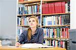 Young female student reading textbook gazing up from library desk