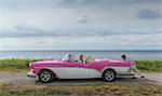 Young man pushing vintage convertible at coast whilst girlfriend drives, Havana, Cuba