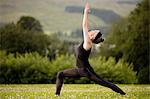 Mature woman practicing yoga warrior pose with arms raised in field