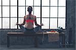 As the city bustles below, this fit, athletic woman sits quietly meditating in lotus position on a wooden bench.