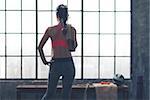 An athletic, strong woman is standing looking out the window in a city loft gym. A towel is thrown over her shoulder, and her hand is on her hip.