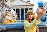 A happy brunette holds up her mobile phone and is smiling while taking a selfie with Rome's Pantheon fountain and Pantheon behind her.