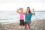 A happy young mother and daughter wearing workout gear are together on the beach at dusk. The mother is kneeling down next to her daughter, and both are flexing their arms to show how strong they are.