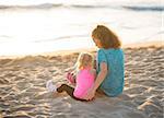 A young mother is sitting next to her daughter, listening to her and playing with her in the sand. Seen from behind, they are both wearing workout gear and are resting, taking time to enjoy the sunset