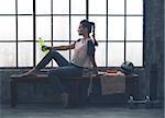 An athletic woman wearing workout gear is sitting relaxing on a wooden bench in a loft gym. Having just finished her yoga workout, she is relaxing and holding a bottle of water.