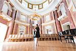 pretty brunette girl with evening dress and heels posing with happy expression in elegant concert hall near ancient empty seat and piano