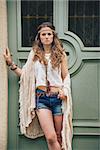 Portrait of brunette hippy-looking woman in jeans shorts, knitted shawl and white blouse standing outdoors against wooden door in old town