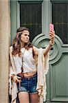 Portrait of hippie woman in boho chic clothes standing outdoors against wooden door in old town and making selfie
