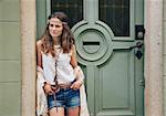 Outdoor fashion portrait of trendy hipster young woman in jeans shorts, knitted shawl and white blouse standing outdoors against olive wooden door in old town