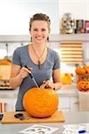 Cheerful modern woman carving a big orange pumpkin Jack-O-Lantern for Halloween party in kitchen. Traditional autumn holiday