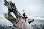 Mature woman practicing yoga lotus position on large driftwood tree trunk at beach