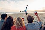 Group of friends sitting on beach, watching friend do cartwheels on and, sunset, rear view