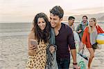 Group of friends walking along beach, young couple looking at smartphone