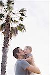 Man standing by a palm tree, carrying his young son in his arms, kissing him on the cheek.