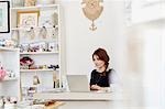 A woman sitting at a desk in a small gift shop, doing the paperwork, managing the business, using a laptop.