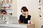 A woman sitting at a desk in a gift shop using a laptop and making a call.