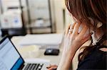 A woman seated in a shop using her laptop and making a phone call.