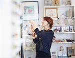 A mature woman in a gift and jewellery shop, looking closely at an object, a shopper or a business owner and jeweller.