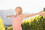 Smiling blonde woman standing in front of grapevine with eyes closed and arms outstretched