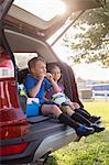 Boy and younger sister sitting in car boot eating oranges on football practice break